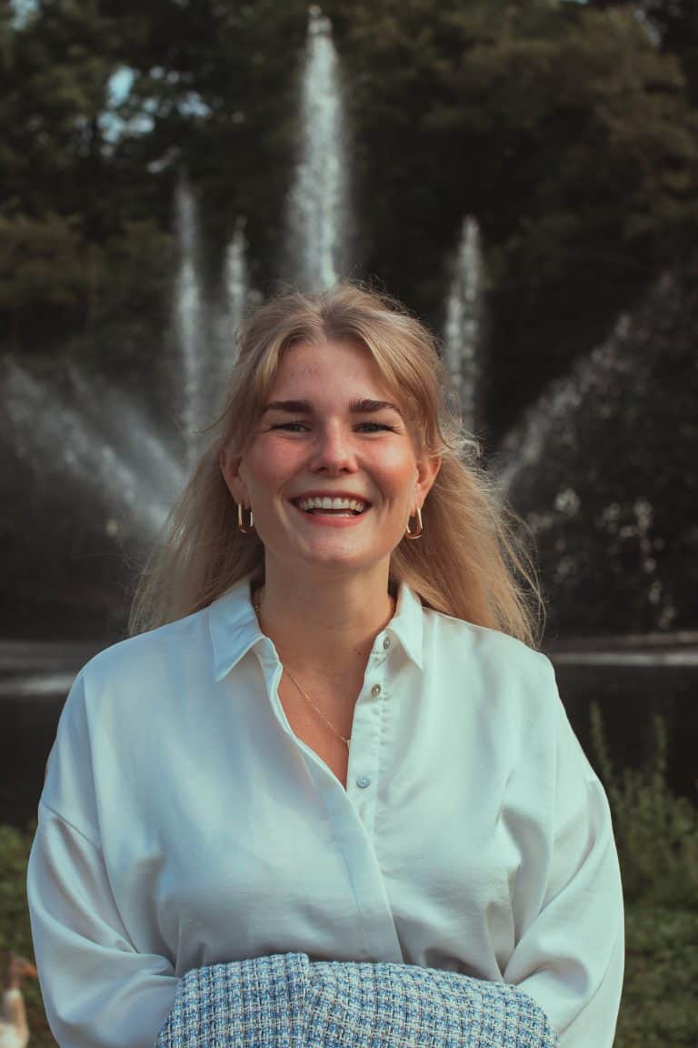 Vrouw die glimlacht in de natuur, met een fontein op de achtergrond, voor werkplezier en groei, welzijn en persoonlijke ontwikkeling op de werkvloer.